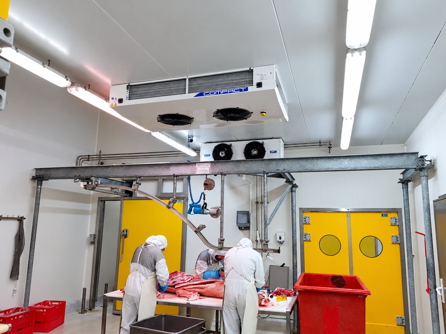 Dans un atelier de découpe réfrigéré, des ouvriers en tenue blanche travaillent de la viande sous des évaporateurs Compact fixés au plafond, près de portes jaunes.