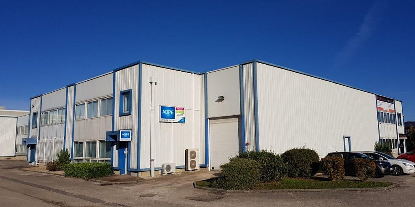 Façade d'un bâtiment industriel à bardage blanc et bleu portant l'enseigne ADPE, avec des arbustes et des voitures garées au premier plan.