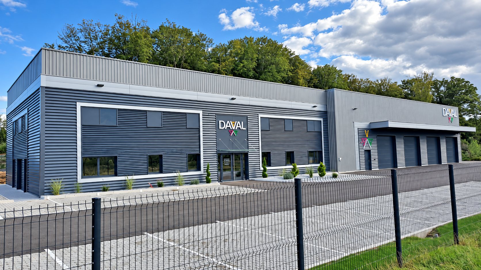 Façade d'un bâtiment industriel moderne en bardage métallique gris portant l'enseigne Daval, avec portes de garage, parking pavé et clôture au premier plan.