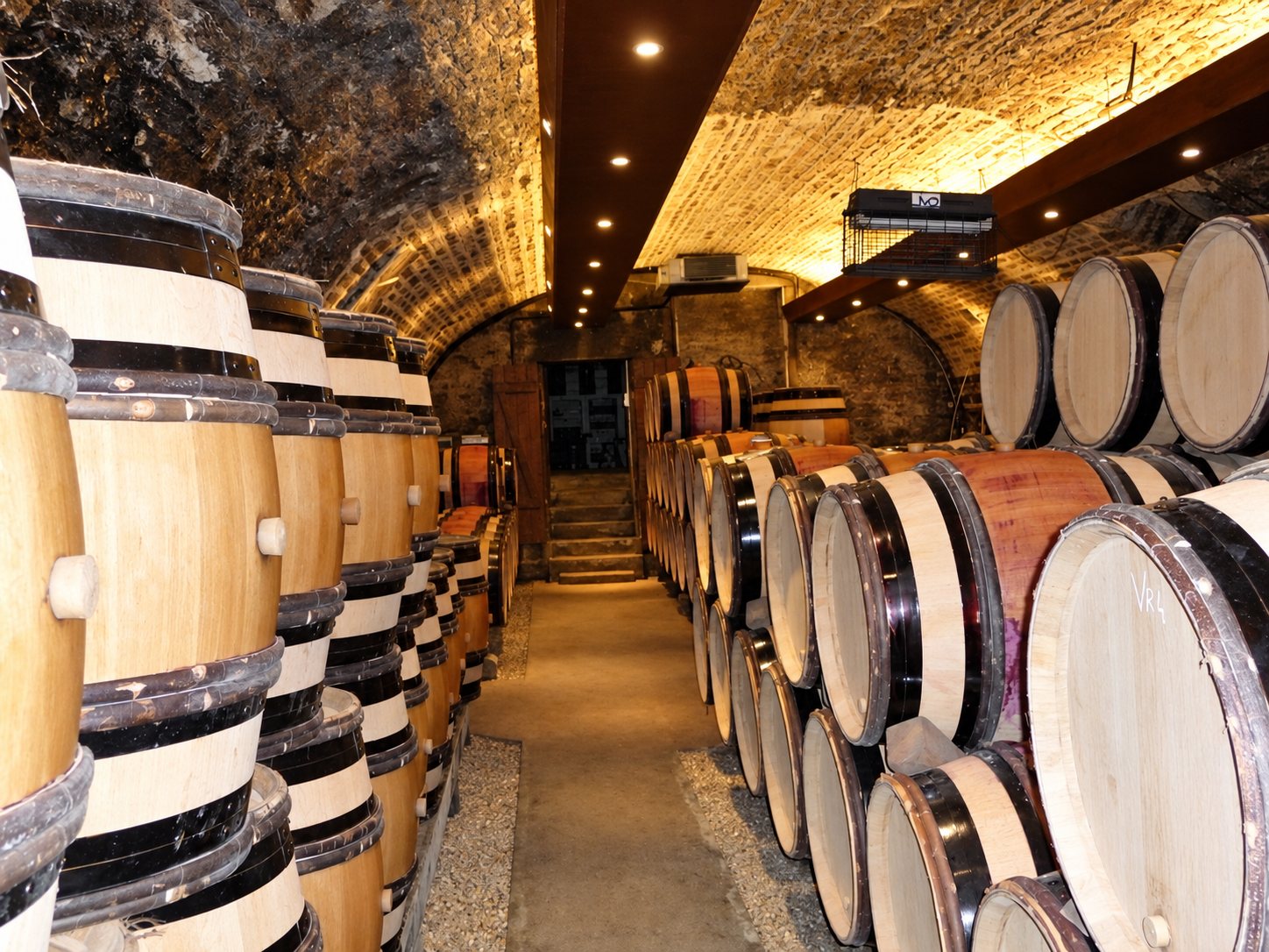 Une cave voûtée en pierre éclairée par des spots, avec des fûts de chêne empilés de part et d'autre d'une allée centrale.
