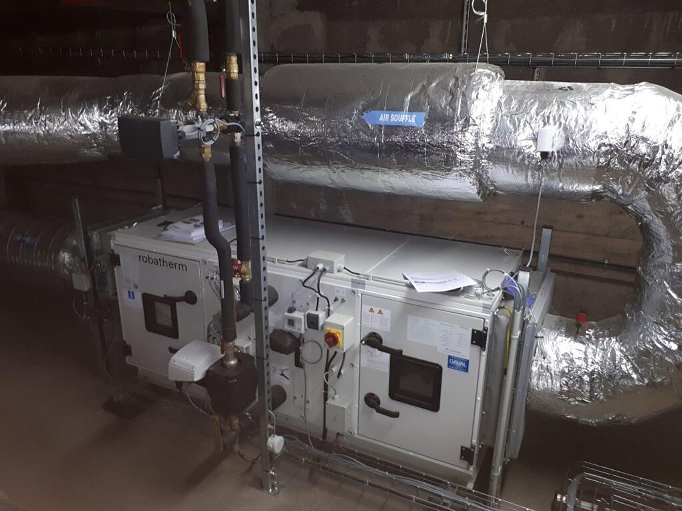Une centrale de traitement d'air Robatherm raccordée à des tuyaux et à des gaines calorifugées en aluminium dans un local technique.