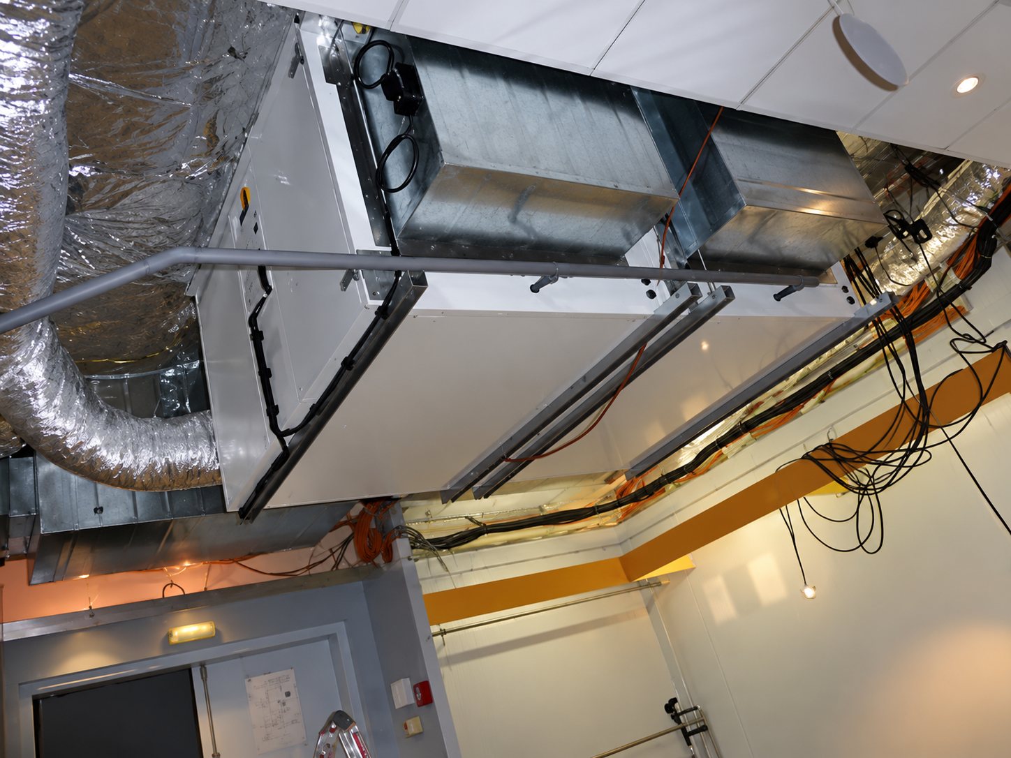 Une centrale de traitement d'air est suspendue sous le plafond d'un local technique, entourée de gaines de ventilation isolées et de câbles.