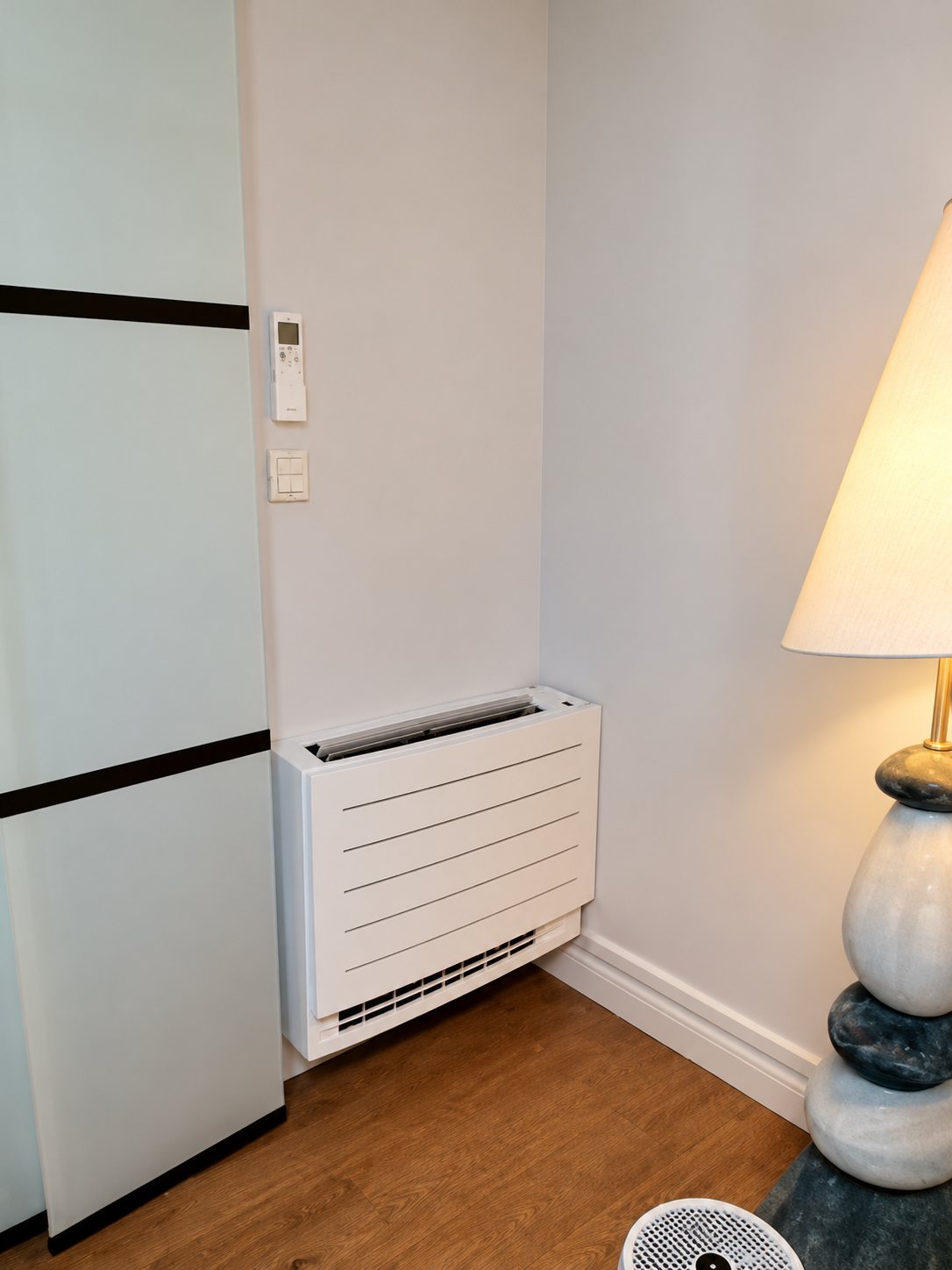 Un climatiseur blanc de type console, posé en bas d'un mur près d'une armoire blanche et d'un pied de lampe, sur un parquet.