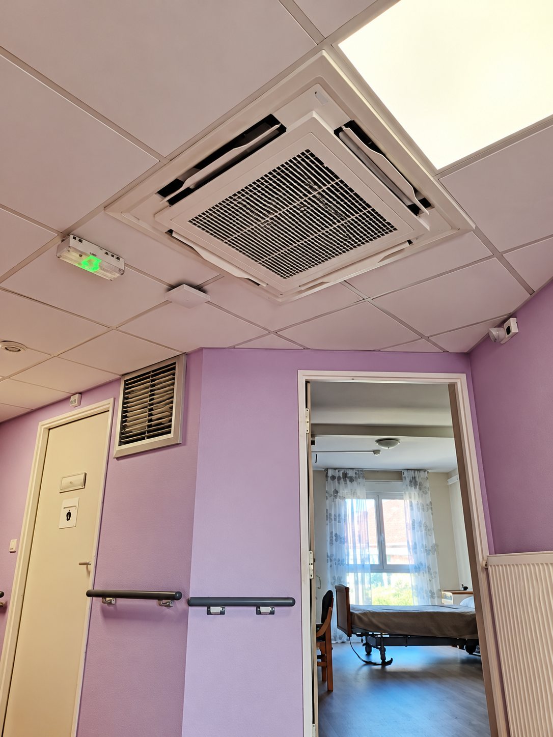 Un climatiseur de type cassette est installé au plafond d'un couloir aux murs violets, avec une chambre médicalisée visible par la porte ouverte.