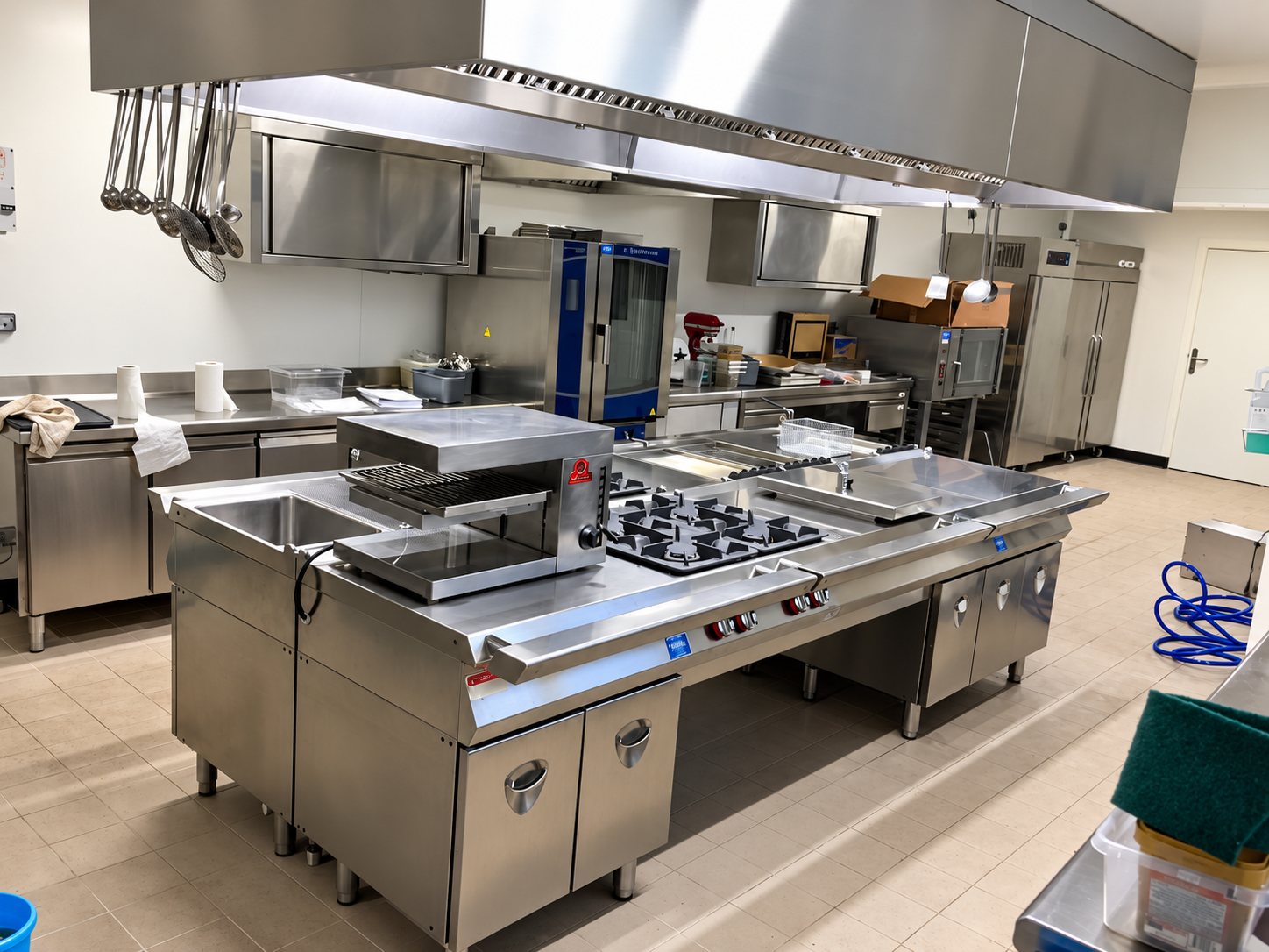 Cuisine professionnelle en inox avec un grand îlot de cuisson central comprenant des feux vifs, une salamandre et une friteuse, sous une hotte aspirante.