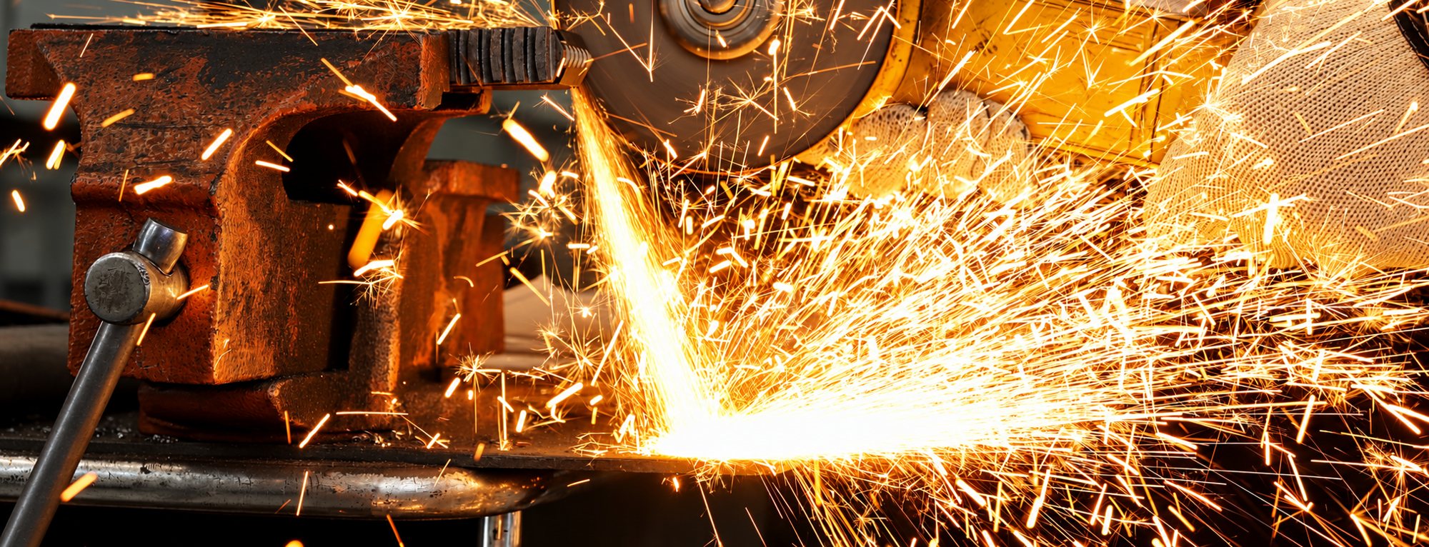 Une meuleuse d'angle découpe une pièce métallique fixée dans un étau rouillé, projetant une gerbe d'étincelles orange dans l'atelier.