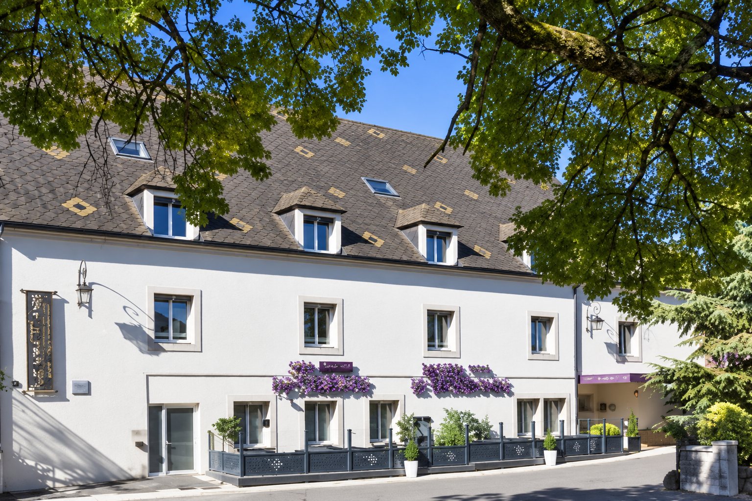 Façade d'un bâtiment blanc à toit d'ardoise avec lucarnes et auvents violets, vue à travers le feuillage d'arbres verts.