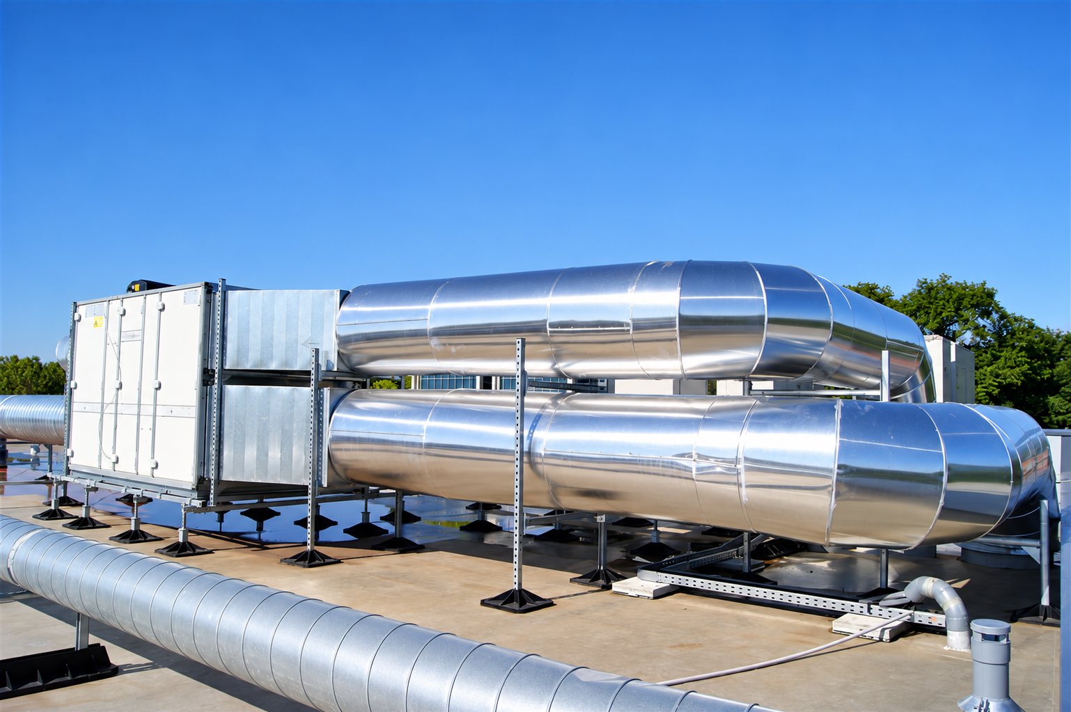 Grosses gaines de ventilation en aluminium brillant reliées à une centrale de traitement d'air installée sur le toit-terrasse d'un bâtiment sous un ciel bleu.