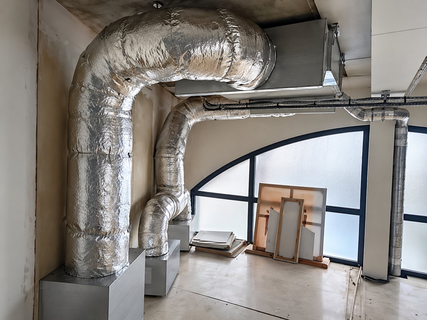 Des gaines de ventilation calorifugées en aluminium courent au plafond et descendent vers des caissons, près d'une grande fenêtre cintrée dans un local en rénovation.