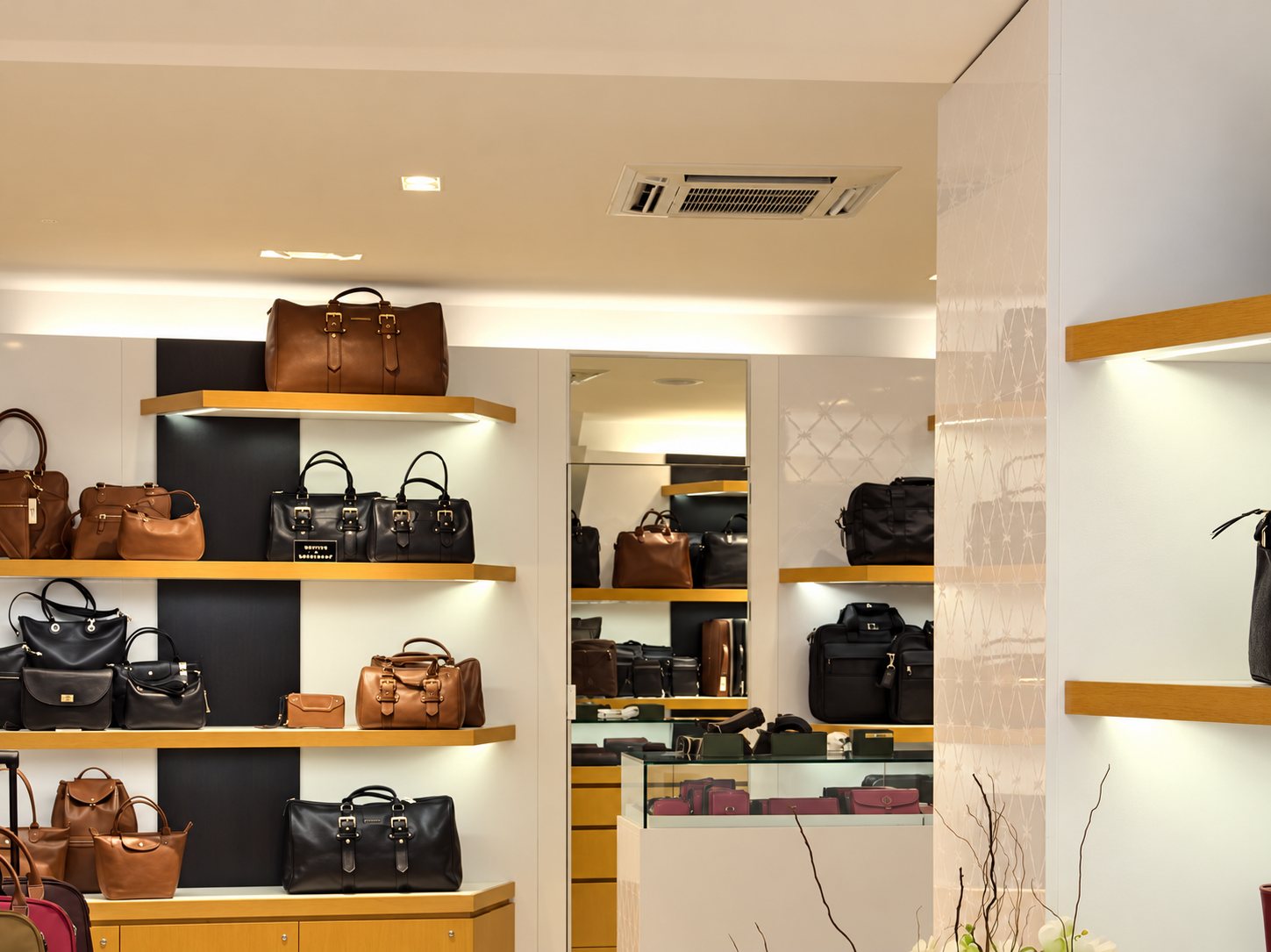 Intérieur lumineux d'une boutique de maroquinerie où des sacs en cuir marron et noir sont présentés sur des étagères en bois, avec une vitrine et un miroir au fond.