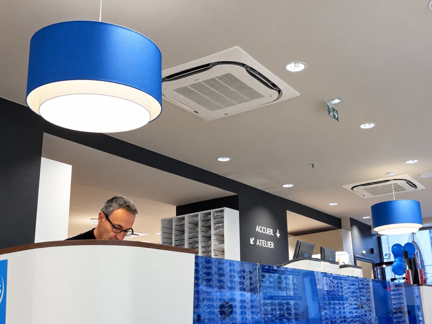 Intérieur lumineux d'un magasin d'optique avec de grands luminaires bleus suspendus, un panneau indiquant accueil et atelier, et un homme à lunettes derrière un comptoir.