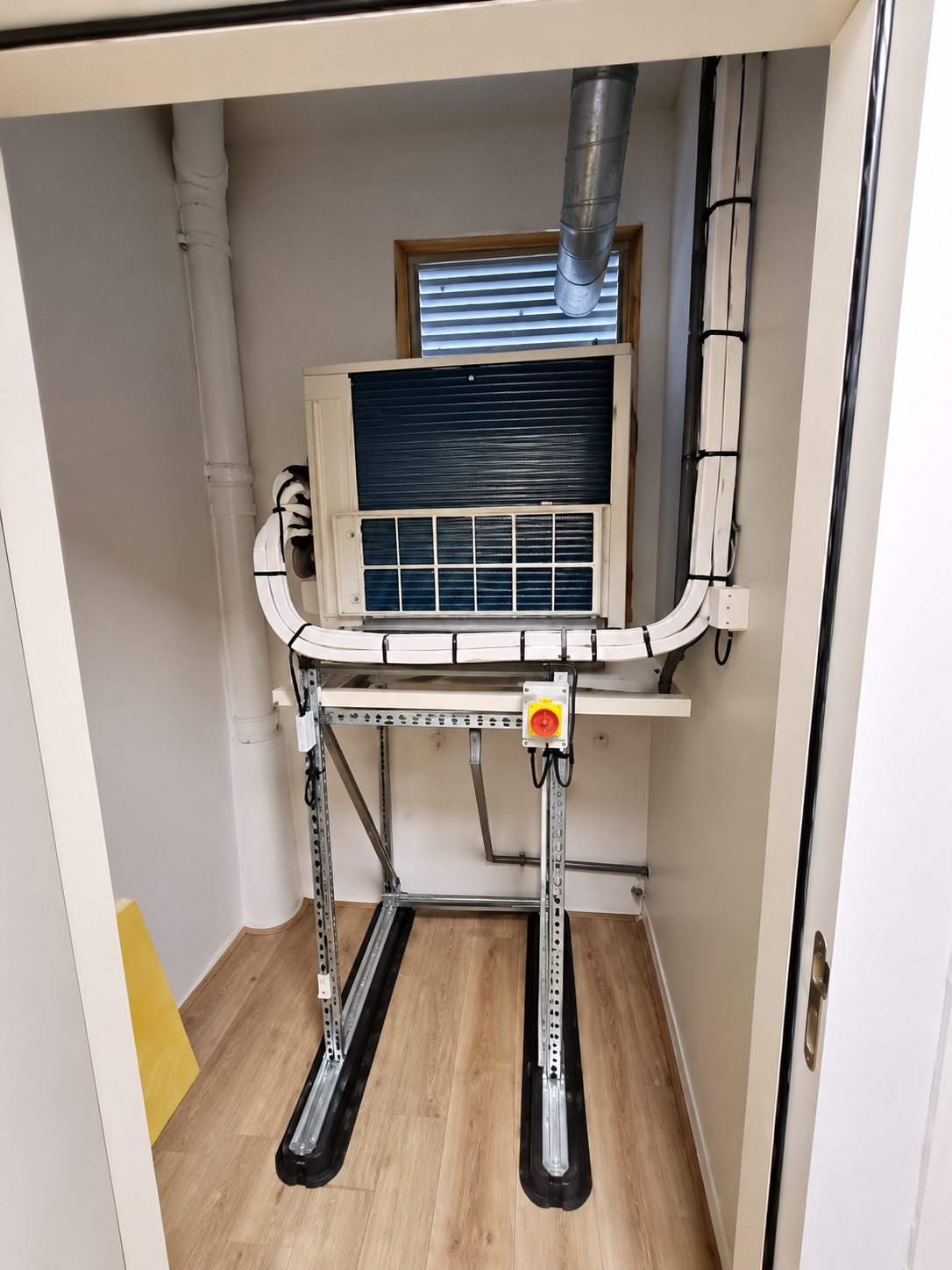 Intérieur d'un petit local au parquet clair contenant un appareil fixé au mur et un support métallique sur rails noirs.