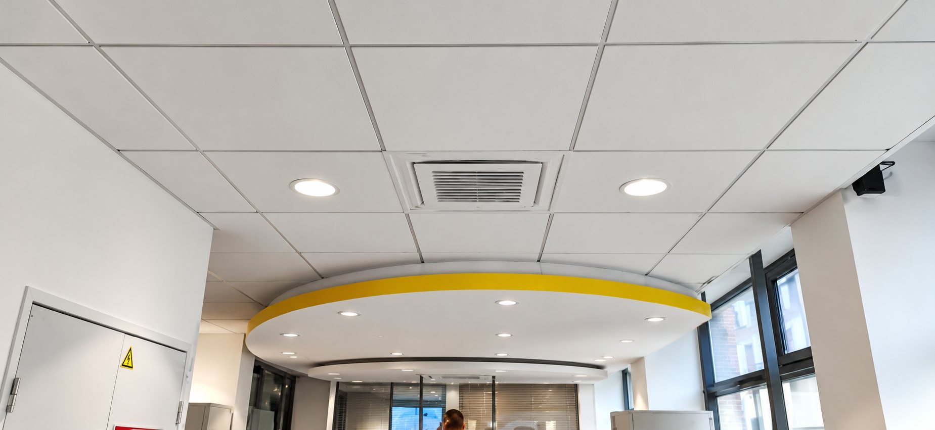 Vue vers le plafond d'un intérieur moderne avec dalles blanches, spots encastrés, une climatisation encastrée et un faux plafond courbe souligné de jaune.