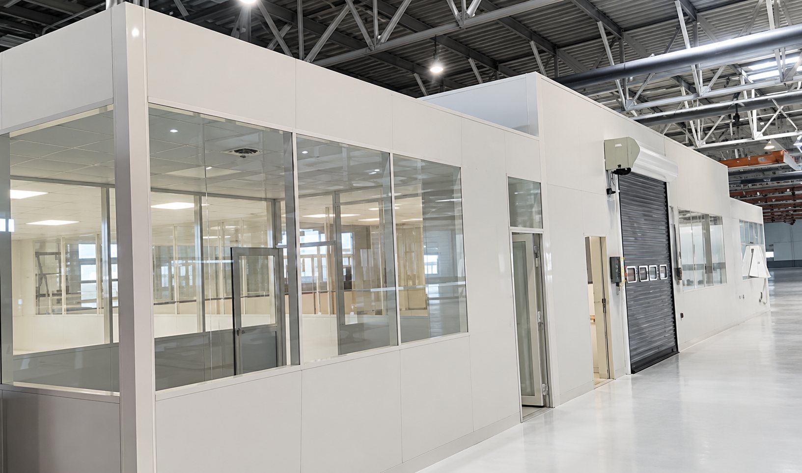 Une structure modulaire blanche aux grandes vitres est installée dans un vaste hangar industriel au sol clair et brillant.