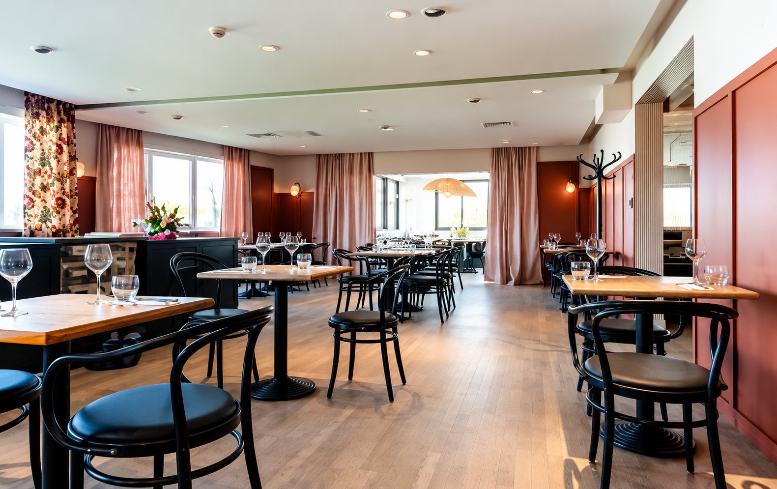 Intérieur d'une salle de restaurant spacieuse avec des tables rondes en bois, des chaises noires, des verres sur les tables et de grandes fenêtres avec rideaux.