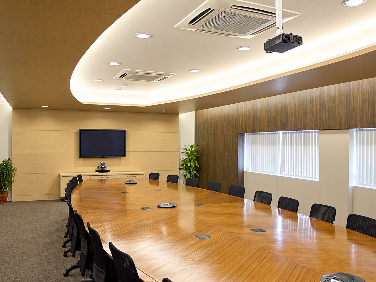 Une salle de réunion lumineuse avec une grande table ovale en bois poli, des fauteuils noirs, un écran mural et un vidéoprojecteur au plafond.
