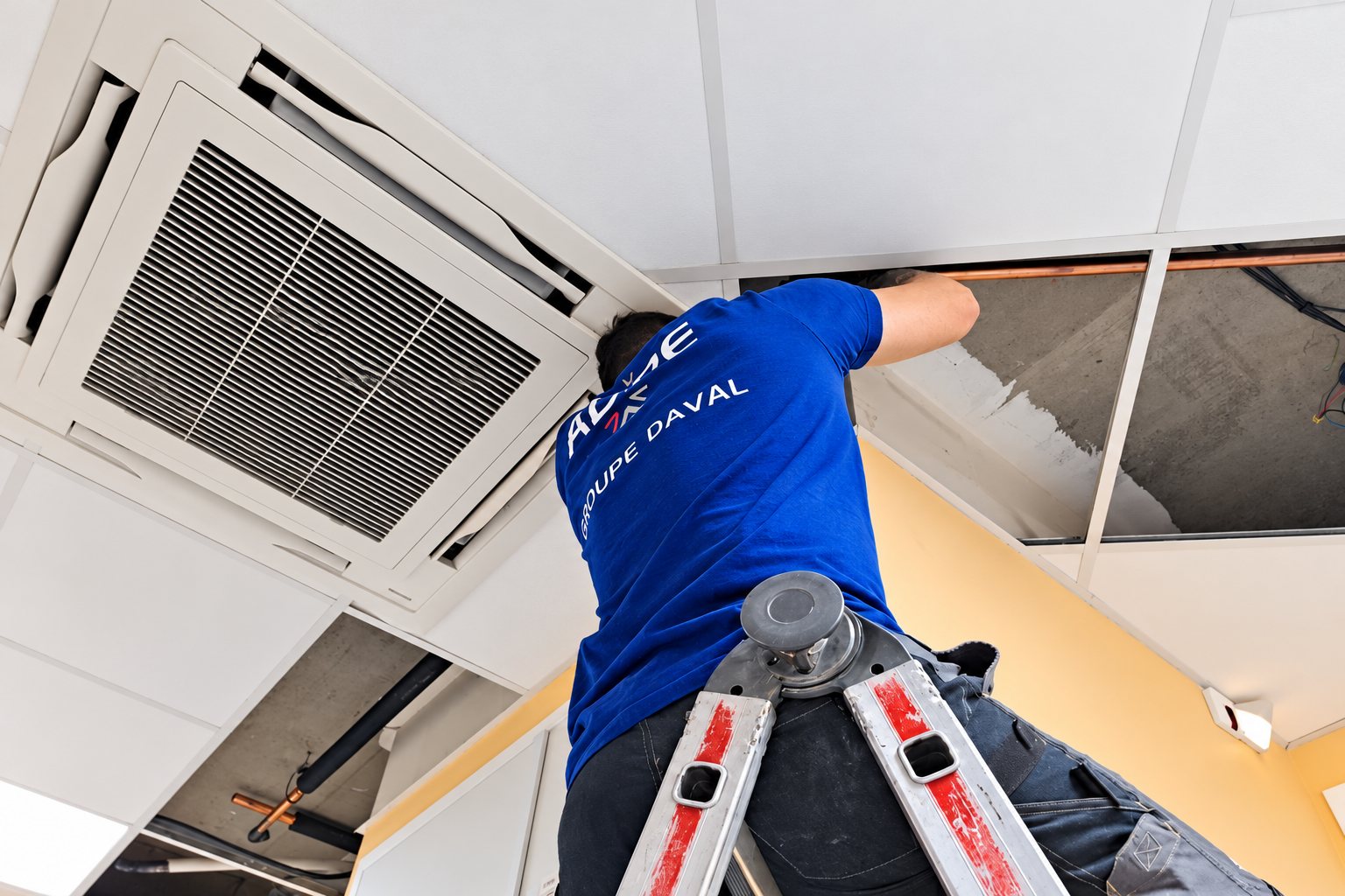 Un technicien en t-shirt bleu, monté sur un escabeau, intervient dans le faux plafond à côté d'une climatisation de type cassette.