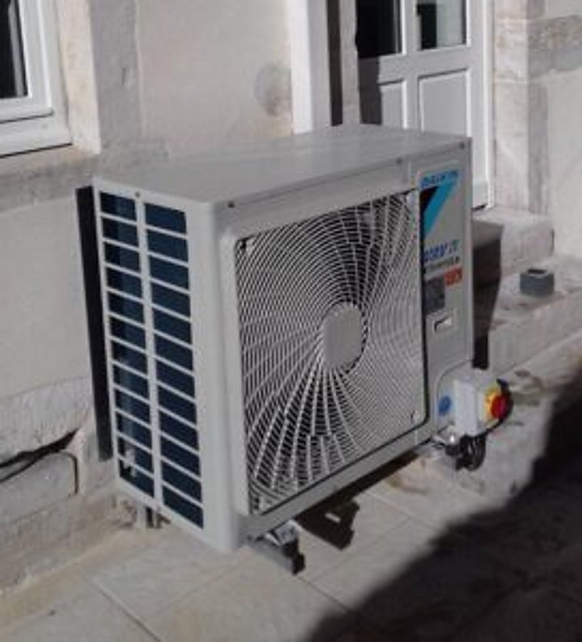 Une unité extérieure de pompe à chaleur blanche est fixée au pied d'un mur en pierre, près d'une porte, sur un sol dallé.